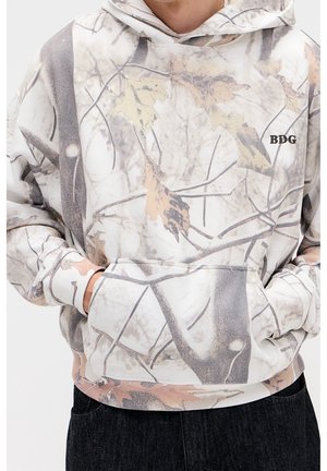 Personne portant un sweat à capuche camouflage pâle avec des motifs de feuilles et de branches, mains dans la poche avant, et "BDG" brodé sur la poitrine.