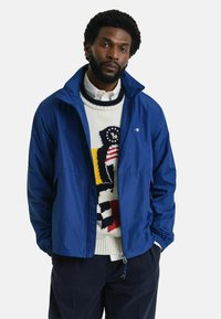 Chaqueta cortavientos azul con cremallera en el frente, material ligero, puños elásticos, llevada sobre un suéter estampado. Diseño limpio, sin logotipos visibles.