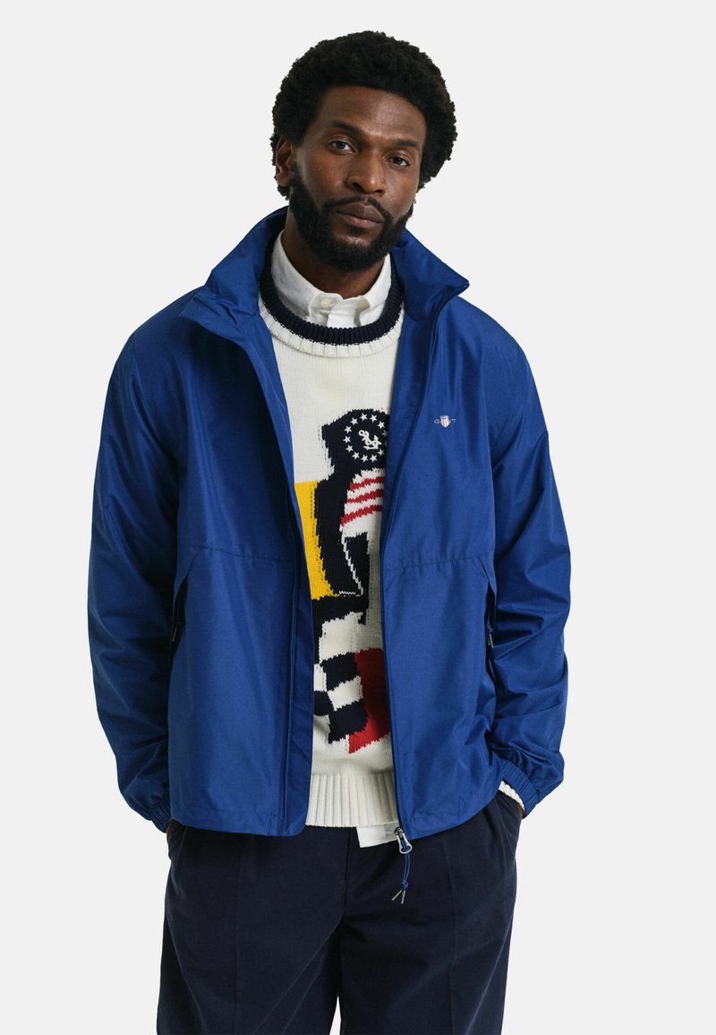 Chaqueta cortavientos azul con cremallera en el frente, material ligero, puños elásticos, llevada sobre un suéter estampado. Diseño limpio, sin logotipos visibles.
