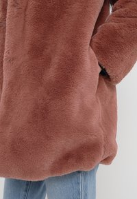 Abrigo de piel sintética marrón con un acabado suave y texturizado, que incluye bolsillos laterales y un ajuste holgado, combinado con jeans de mezclilla azul claro.