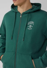 Grüne Zip-Hoodie mit Kordelzugkapuze, Fronttaschen und einem weißen Logo-Aufdruck auf der linken Brust. Weicher Stoff mit glatter Textur.