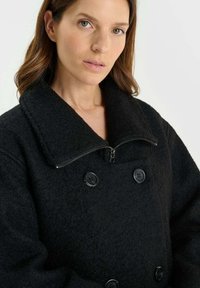 Zwarte wollen jas met een dubbelzijdig ontwerp, grote kraag, rits en twee knopen. Zachte, gestructureerde stof met een relaxte pasvorm.