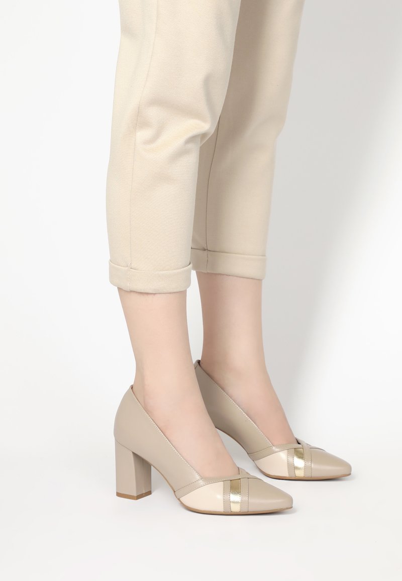 Béžové lodičky na vysokém podpatku s špičatou špičkou, vyrobené z lesklého materiálu a se zlatým akcentem na přední straně. Design se silným podpatkem.