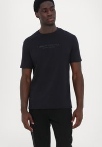Juodas medvilnės marškinėliai trumpomis rankovėmis, su subtiliu iškilniu logotipu "Armani Exchange Milano/New York" priekyje.