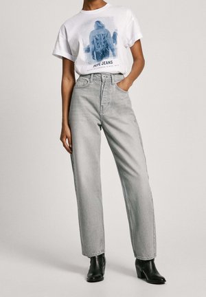 Henkilö, joka pukeutunut vaaleanharmaisiin korkeavyötäröisiin farkkuihin, mustiin nilkkureihin ja valkoiseen lyhythihaiseen T-paitaan, jossa on sininen grafiikka ja teksti "PEPE JEANS."