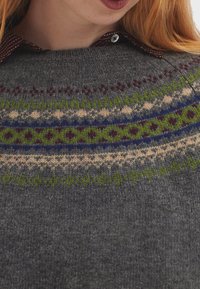 Pull en tricot gris avec un col rond, arborant des motifs Fair Isle multicolores en vert, bleu, marron et crème sur la poitrine.