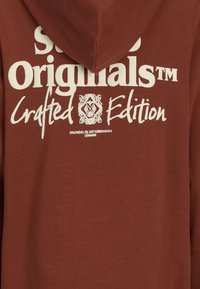 Hnedá mikina s kapucňou na stiahnutie. Obsahuje biely potlač s nápisom "Super Originals™ Crafted Edition" a zložitými dizajnovými detailmi. Mäkká tkanina.