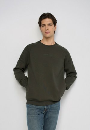 Hombre con sudadera acolchada de color verde oscuro con el logo "Salsa Jeans" y vaqueros azules, de pie frente a un fondo claro liso con las manos en los bolsillos.