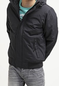 Giubbotto imbottito nero con cappuccio, presenta una chiusura frontale con zip e tasche laterali, polsini elastici e una texture liscia. Indossato sopra una maglia blu a petrolio.