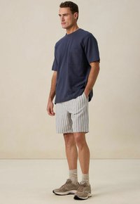 T-shirt bleu marine à manches courtes avec une poche poitrine, associé à un short à rayures blanches et en couleurs pastel. Baskets beiges et chaussettes grises.