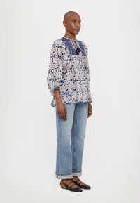 Bluse mit Blumendruck in Weiß, Blau und Rot, mit Schlüsselloch-Ausschnitt und gerafften Ärmeln, kombiniert mit hellblauen, geraden Jeans und dunklen Ballerinas.