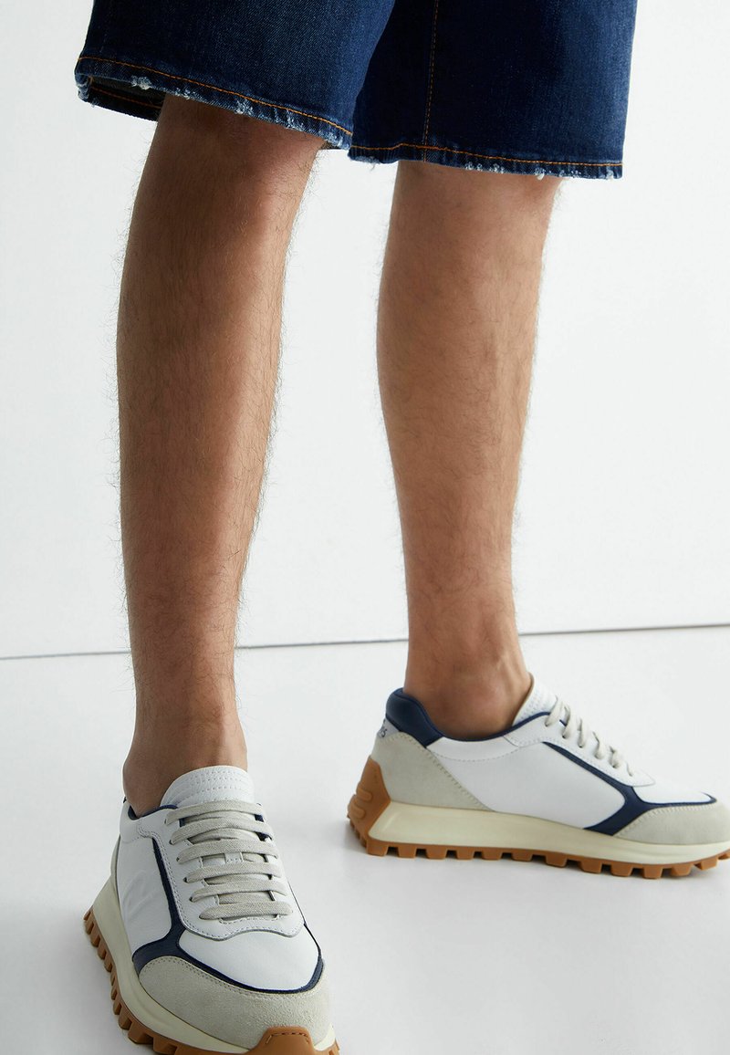 Scarpe da ginnastica bianche e blu navy con dettagli testurizzati, lacci rotondi e suola spessa in gomma. Indossate con pantaloni corti di jeans scuri.