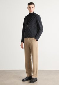 Black zip-up jacket with hood and smooth texture, paired with light beige trousers. Model wears black ankle boots, standing on a neutral background.