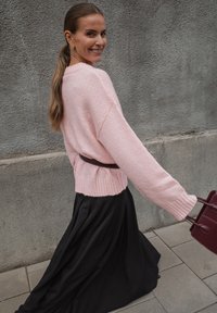 Sieviete ar garu asti, ģērbusies rozā džemperī un melnās svārkos, staigā ārā pa flīzētām ielām ar teksturētu pelēku sienu fonā.