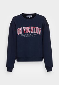 Marinblå crewneck-tröja med "ON VACATION" i stora röda bokstäver och "Stolt medlem av Do Nothing Club" i mindre vita tecken nedanför.
