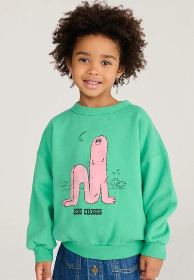 Bambino con capelli ricci che indossa una felpa verde con un serpente rosa da cartone animato e il testo "BOBO CHOSES", in piedi davanti a uno sfondo semplice.