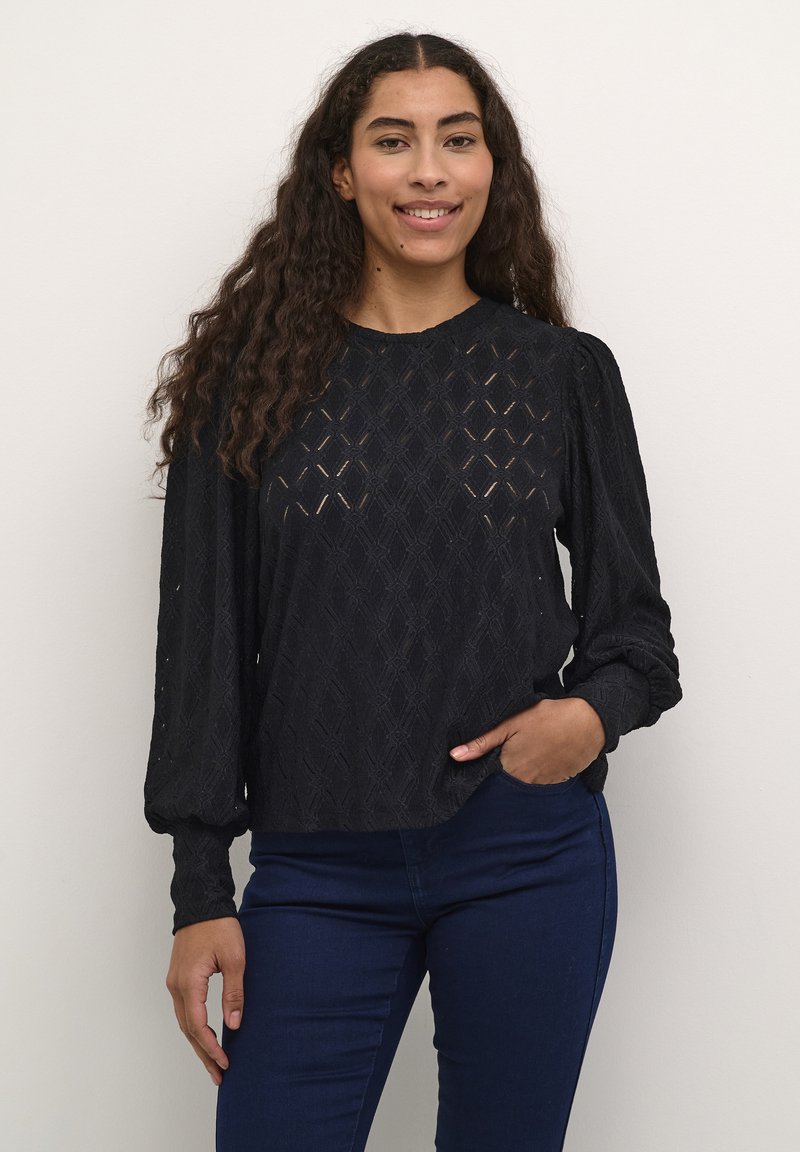 Svart långärmad topp med strukturerade diamantmönster, rund halsringning och puffärmar, kombinerad med mörkblå tajta jeans.