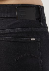 Acercamiento a un bolsillo trasero y la cinturilla de unos vaqueros de mezclilla negros, con una etiqueta de marca G-Star RAW de cuero negro y una pequeña etiqueta metálica RAW en el bolsillo.