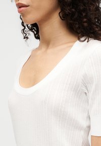 Haut blanc à manches courtes en tissu tricoté texturé avec un col en U et un motif côtelé vertical.