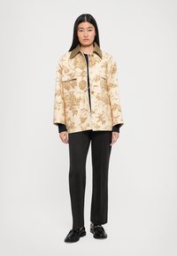 Beige Blazer mit Blumenmuster und Kragen, mit zwei vorderen Taschen, über einem schwarzen Oberteil getragen und kombiniert mit schwarzen Hosen und Schuhen.