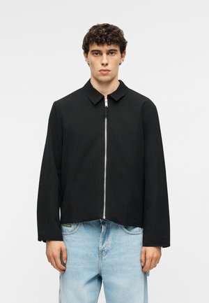 Jeune homme aux cheveux bouclés portant une veste noire zippée et un jean bleu clair, debout devant un fond blanc uni.