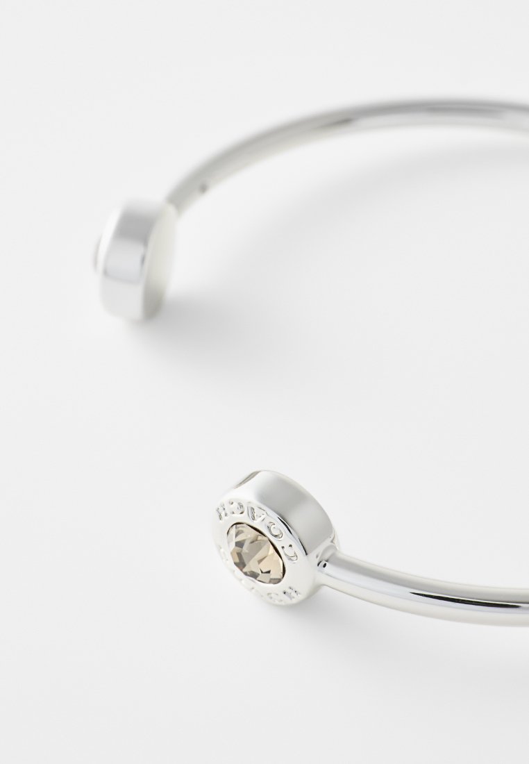 Pulsera de brazalete de plata que cuenta con un acento de gema clara en un extremo y un cierre liso y redondeado en el otro. Diseño simple y moderno.