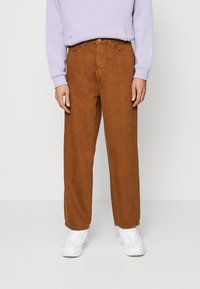 Brązowe szerokie spodnie z miękkiego materiału, z przednimi kieszeniami i metalowym guzikiem w pasie, zestawione z jasnofioletową bluzą i białymi sportowymi butami.