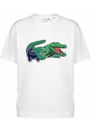 Biała koszulka Lacoste z dużą stylizowaną grafiką krokodyla w kolorach zielonym i granatowym, z otwartą paszczą i ostrymi zębami na przodzie.