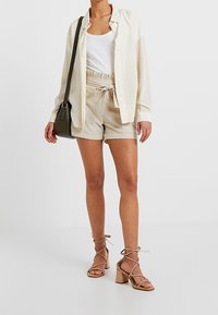 Lekkia w kolorze kości słoniowej koszula zapinana na guziki na białym topie, w połączeniu z beżowymi szortami na sznurku i beżowymi sandałami na obcasie z paskami. Zielona torebka.