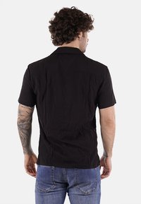 Maglietta nera a maniche corte con colletto, caratterizzata da un tessuto texture. La vista posteriore mostra cuciture piatte e un orlo dritto.