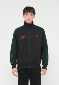 Sort lynlåstrøje med grønne striber langs ærmerne, med rød broderet tekst og et lille rødt logo på brystet.