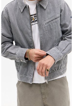 Person iført en blåhvidstribet jakke over en hvid grafisk t-shirt, justerer jakkeærmet med synligt guldarmbånd og ring.