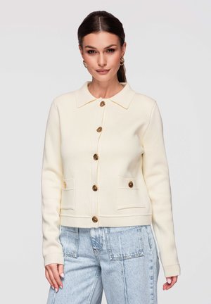 Mujer con cabello oscuro recogido en una cola de caballo que lleva un cárdigan crema abotonado con bolsillos y vaqueros de tiro alto azul claro, de pie contra un fondo liso.