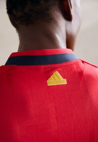 Sarkans sporta krekls ar jūras zila apkakli, ar vertikālām svītrām un dzelteno Adidas logo aizmugurē. Gluda tekstūra ar elpojošu audumu.