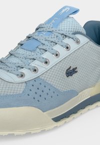Gros plan d'une basket bleu clair et blanche avec des détails en mesh et en daim, des lacets bleus et un logo crocodile sur le côté.