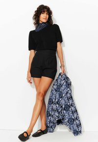 Pull noir à manches courtes, short noir taille haute, veste à motifs bleus et noirs, et ballerines noires à clous.