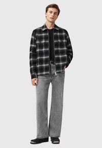 Camicia overshirt a quadri neri e grigi con due tasche sul petto, abbinata a dei jeans grigi larghi e scarpe nere con dettagli bianchi.