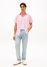 Ανοιχτό ροζ πουκάμισο με κοντά μανίκια και κουμπιά, με λευκό μπλουζάκι από μέσα, συνδυασμένο με ανοιχτό μπλε τζιν και λευκά αθλητικά παπούτσια με καφέ σόλες.