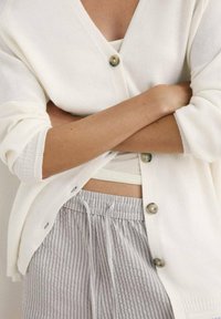 Cardigan bianco lavorato a maglia con scollo a V, dotato di grandi bottoni e maniche morbide, sovrapposto a un top leggero senza maniche e pantaloni grigi a righe con coulisse.