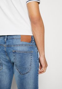 Blue denim jeans with a faded finish, featuring back pockets and a brown leather logo patch labeled "INDIE DUE" at the waistband.