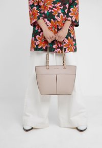 Jasnoróżowa skórzana torba typu tote z podwójnymi uchwytami i akcentującym przeszyciem. Posiada wyraźny kształt i dwie przednie kieszenie.