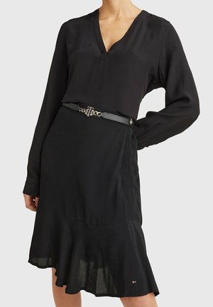 Kobieta w czarnej bluzce z długim rękawem i dekoltem w serek, włożonej do czarnej, falbaniastej spódnicy z czarnym paskiem z metalową klamrą.