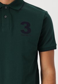 Polo košeľa v tmavozelenej bavlne s vyšitým námorníckym číslom "3" a logom "HACKETT". Klasický golier a kontrastné manžety rukávov.