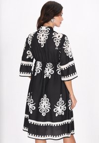 Rochie neagr�ă cu modele florale albe, mâneci trei sferturi, talie strânsă și fustă evazată cu o margine decorativă.