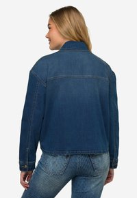 Giacca di jeans di colore blu scuro con un design cropped, con polsini abbottonati e dettagli della cucitura del giromanica sul retro. Indossata sopra jeans a righe blu chiaro.