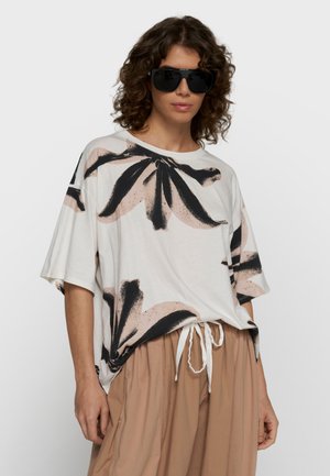 Frau trägt übergroßes weißes T-Shirt mit großem schwarzem und beigem Blumenmuster, beige Kordelzughose und große schwarze Sonnenbrille.