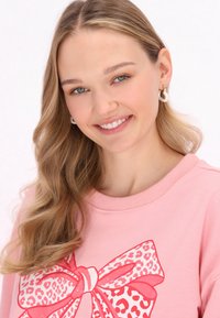 Felpa rosa chiaro con una grande grafica a forma di fiocco leopardato colorato. Il modello mostra capelli lunghi e ondulati e piccole orecchini a cerchio.