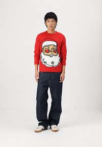 Pull rouge avec un design de Père Noël, affichant une barbe blanche et des détails colorés. Associé à un jean noir décontracté et des chaussures blanches.