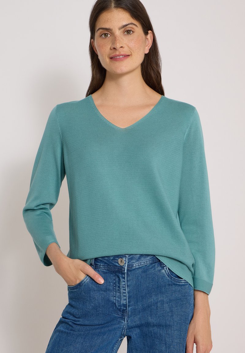 Femme aux longs cheveux foncés portant un haut à manches longues à col en V couleur sarcelle et un jean bleu, une main dans la poche.