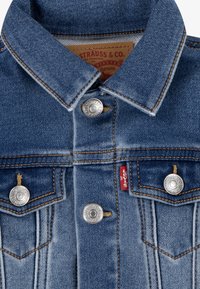 Blå denimjacka med knäppning, silverf�ärgade detaljer, bröstfickor och kontrasterande orange sömmar. Varumärkesetikett synlig inuti.